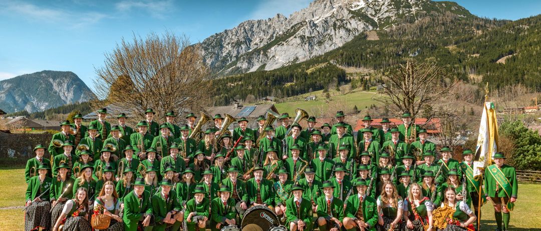 Eine große Gruppe von Menschen in grünen Uniformen und Hüten, möglicherweise eine Marschkapelle, posiert für ein Foto auf einer Wiese mit Bergen im Hintergrund.