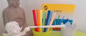 Eine weiße Engelsfigur hält einen Bleistift. Es gibt farbige Stifte in einem grünen Becher, eine Glas-Sanduhr und eine rosa Seife auf einem Teller. Eine Buddha-Statue, ein Buch und ein weißer Spitzenkorb sind ebenfalls vorhanden.