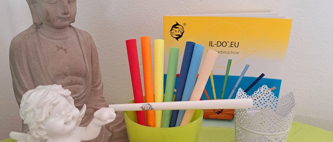 Eine weiße Engelsfigur hält einen Bleistift. Es gibt farbige Stifte in einem grünen Becher, eine Glas-Sanduhr und eine rosa Seife auf einem Teller. Eine Buddha-Statue, ein Buch und ein weißer Spitzenkorb sind ebenfalls vorhanden.