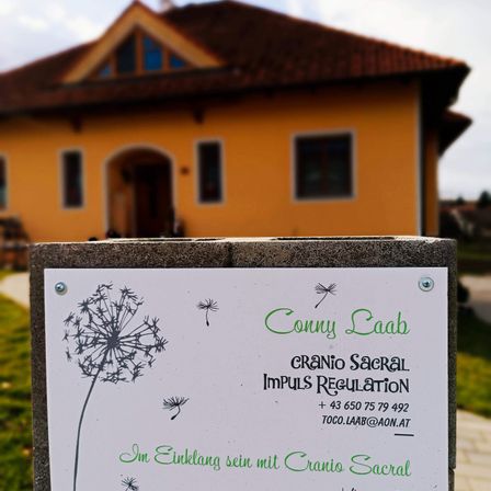 Ein weißes Schild mit einer Löwenzahn-Zeichnung steht vor einem gelben Haus mit einem braunen Dach. Auf dem Schild steht 'Conny Raab Cranio Sacral Impuls Regulation'. Das Haus hat Glasfenster und eine Holztür.