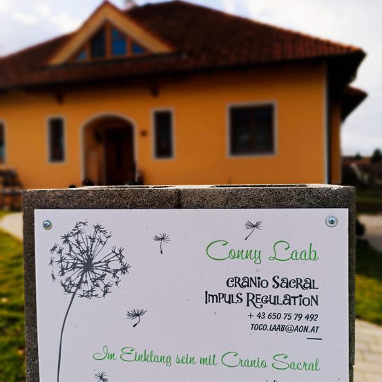 Ein weißes Schild mit einer Löwenzahn-Zeichnung steht vor einem gelben Haus mit einem braunen Dach. Auf dem Schild steht 'Conny Raab Cranio Sacral Impuls Regulation'. Das Haus hat Glasfenster und eine Holztür.
