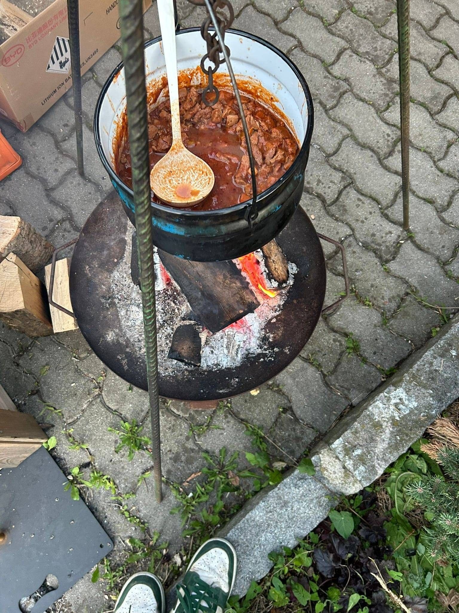Ein Topf mit Eintopf kocht über einem Feuerkorb, mit einem Holzlöffel darauf. Der Topf steht auf einem Metallständer, und der Feuerkorb ist von Holz umgeben.