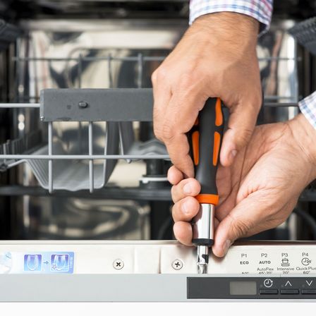Eine Person benutzt einen Schraubendreher, um die Einstellungen einer Spülmaschine zu ändern. Die Spülmaschine hat ein Bedienfeld mit verschiedenen Optionen wie AutoFlex, Intensiv und Quick Plus.