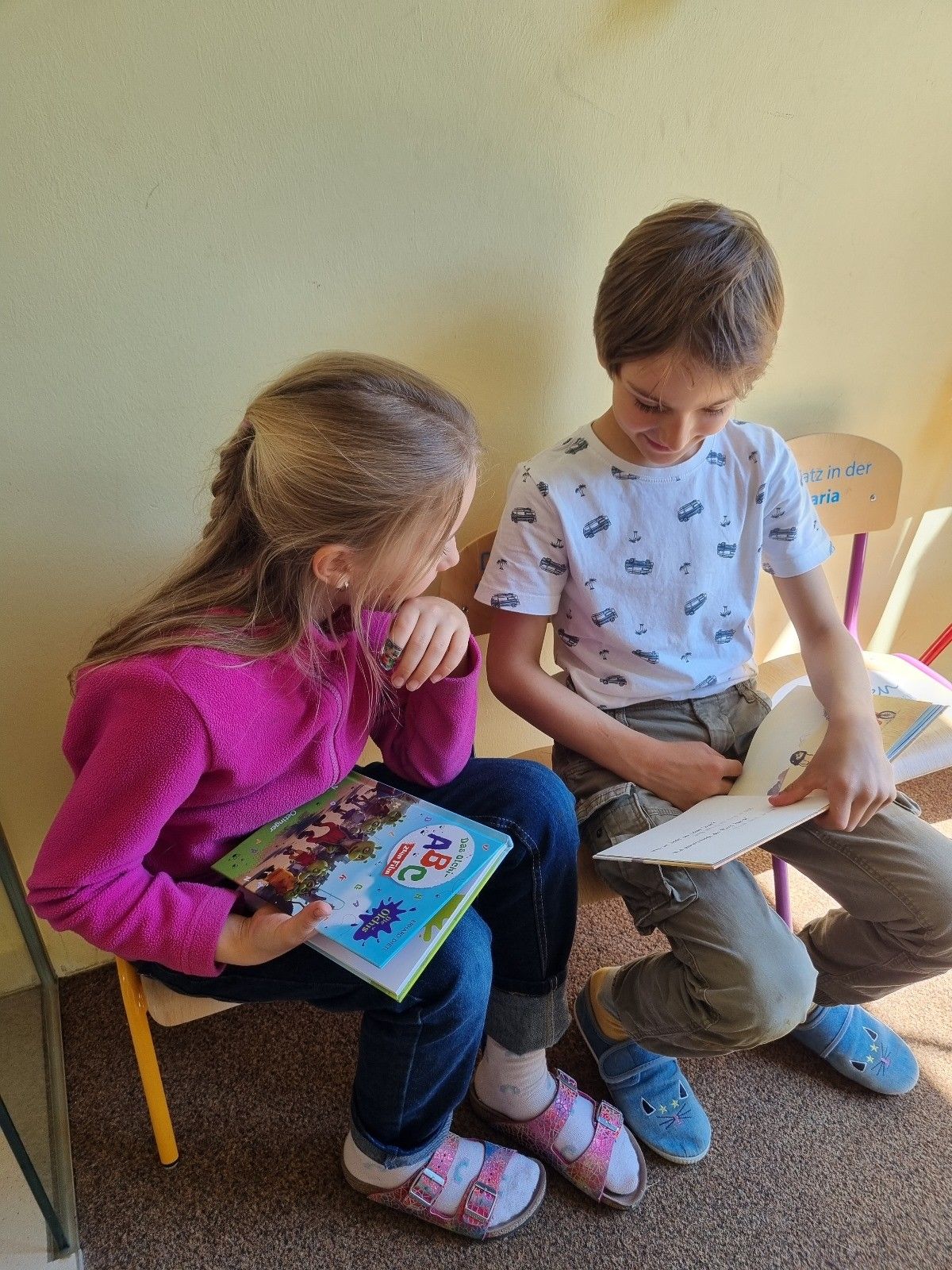 Zwei Kinder sitzen auf Stühlen in einem Klassenzimmer, eines liest ein Buch mit einem bunten Cover und das andere liest ein Buch mit einem weißen Cover. Das Mädchen trägt einen rosa Pullover und der Junge trägt ein weißes T-Shirt mit gedruckten Autos.