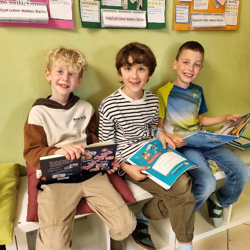 Drei Jungen sitzen auf einer Bank, lächeln und halten Bücher in den Händen. Einer trägt ein 'New York'-Sweatshirt, ein anderer ein gestreiftes Hemd und der dritte ein blau-gelbes Hemd. Sie lesen Bücher mit bunten Einbänden.