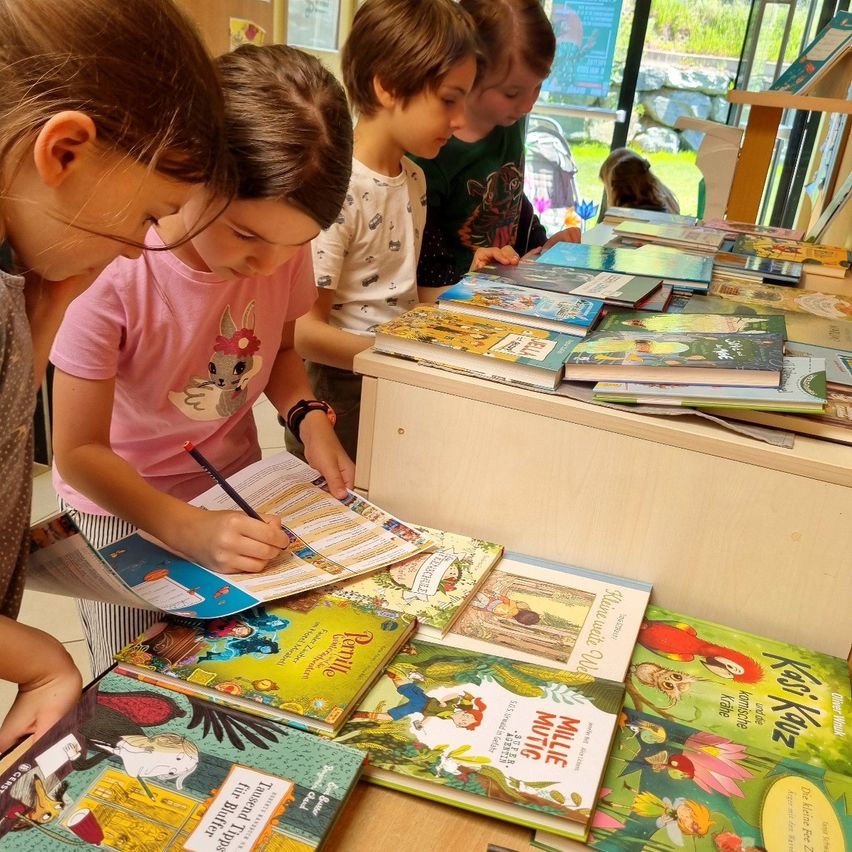Mehrere Kinder stehen um einen Tisch mit Büchern herum. Ein Mädchen in einem rosa T-Shirt schreibt mit einem Stift auf ein Papier. Andere Bücher sind auf dem Tisch gestapelt.