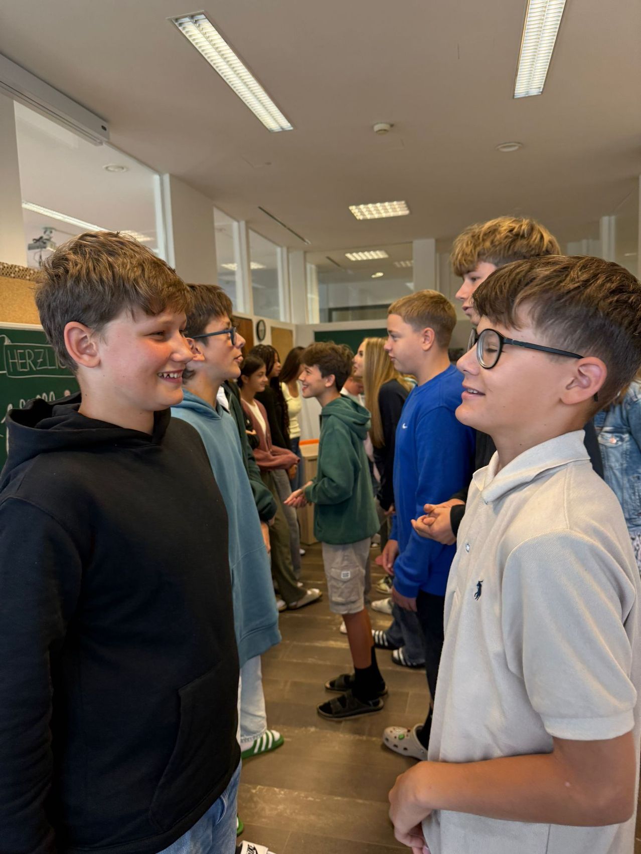 Eine Gruppe von Schülern, Jungen und Mädchen, versammelt sich in einem Flur, wahrscheinlich in einer Schule. Sie tragen Freizeitkleidung, und einige tragen Brillen. Die Schüler schauen auf etwas auf der rechten Seite.