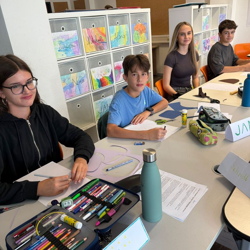 Vier Personen sitzen um einen langen Tisch herum, wahrscheinlich in einem Kunstunterricht. Vor ihnen liegen verschiedene Malutensilien und Papiere. Die Frau links trägt eine Brille.