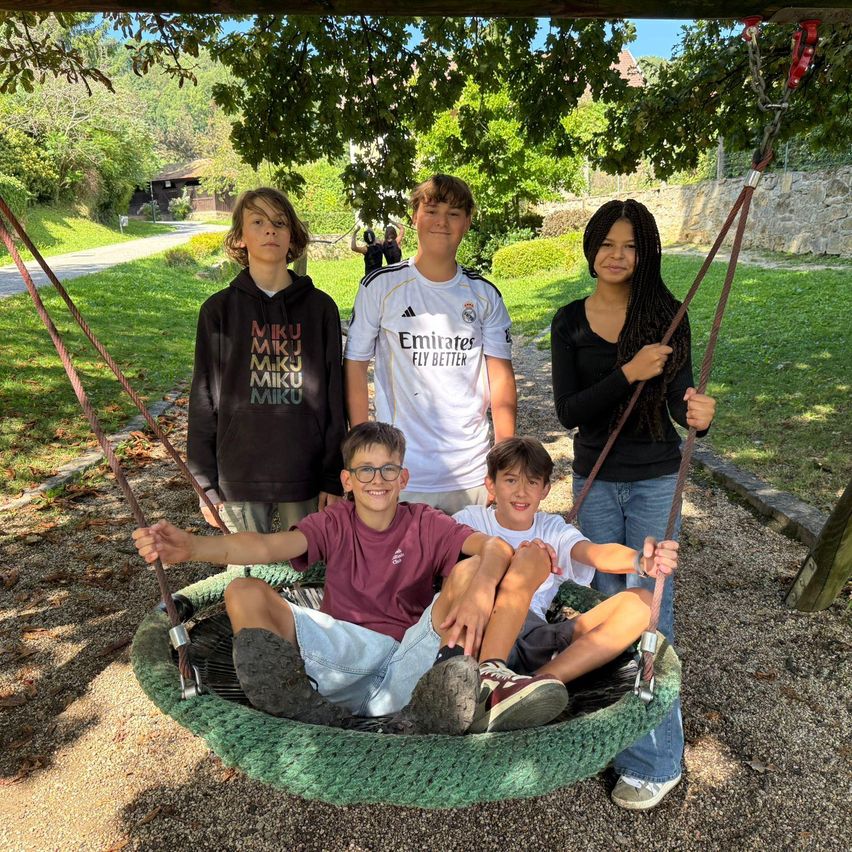 Fünf junge Menschen stehen um eine Reifenschaukel in einem Park herum. Zwei sitzen im Reifen, einer steht dahinter. Einer trägt eine Kapuzenjacke, ein anderer ein Shirt mit 'Miku' darauf.