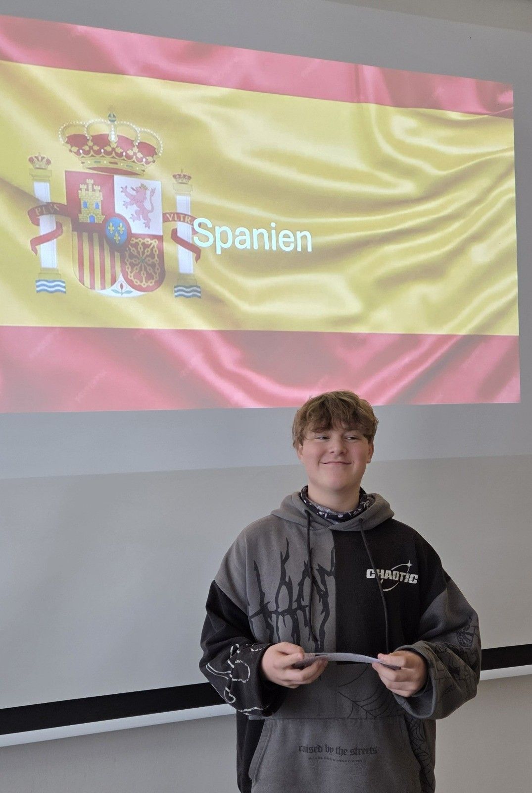 A young boy in a hoodie stands in front of a Spanish flag backdrop, smiling.
