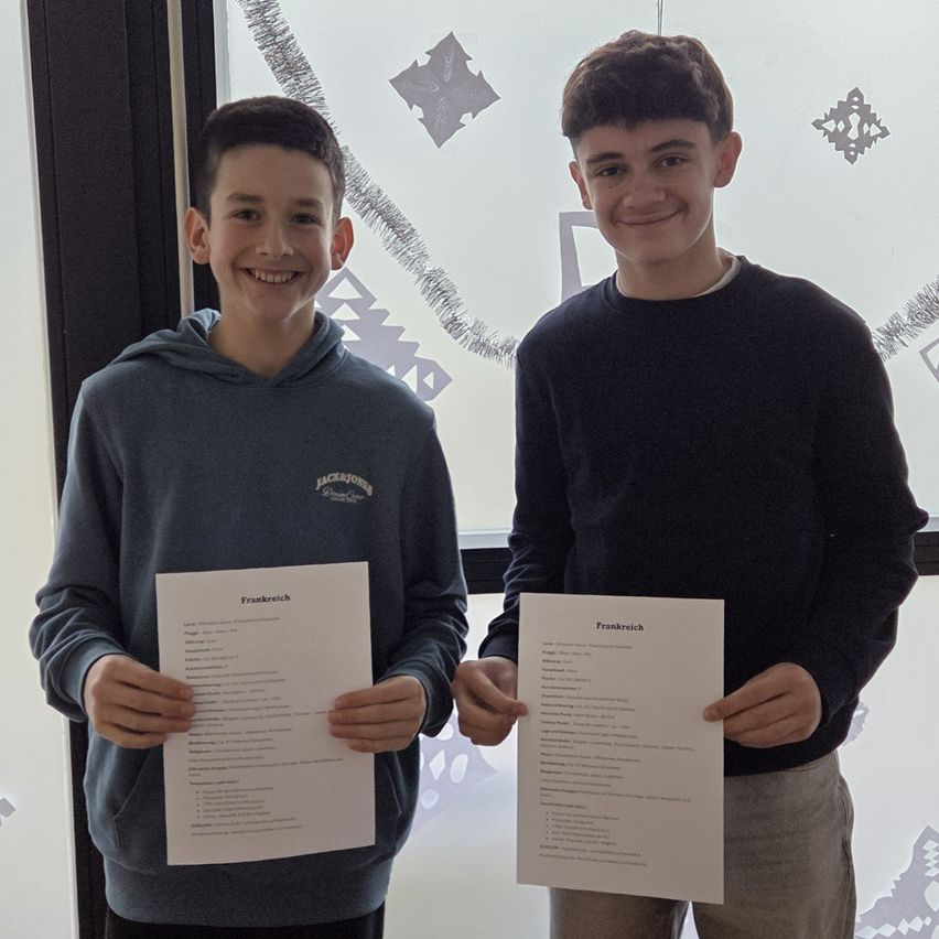 Two boys holding papers, one smiling, both looking at the camera. The boy on the left wears a blue hoodie, and the boy on the right wears a black shirt. They stand before a decorated window.