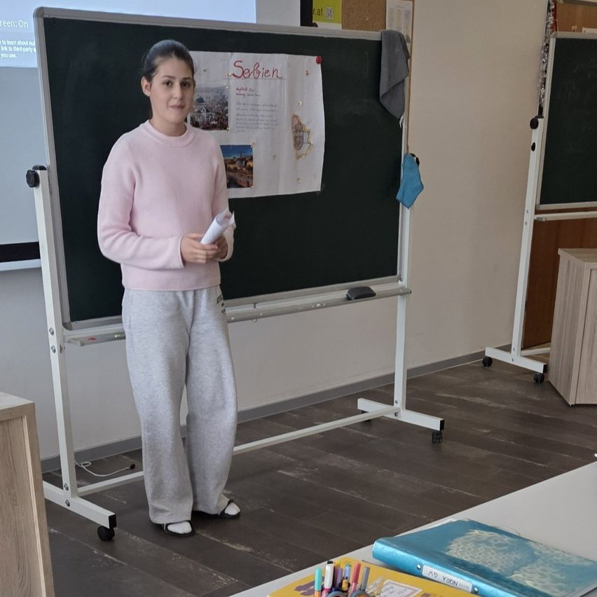 A girl in a pink sweater and gray pants stands in front of a blackboard, holding a paper. Behind her, a whiteboard is mounted on a stand with a projector screen above. Papers are pinned to the board. A blue cloth hangs on the right side.