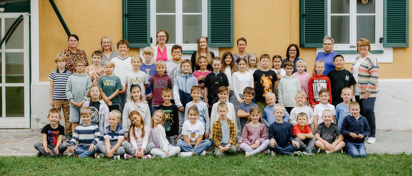 Eine Gruppe von Kindern und Lehrern posiert für ein Foto vor einem gelben Gebäude mit zwei Fenstern und grünen Fensterläden.