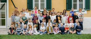 Eine Gruppe von Kindern und Lehrern posiert für ein Foto vor einem gelben Gebäude mit zwei Fenstern und grünen Fensterläden.