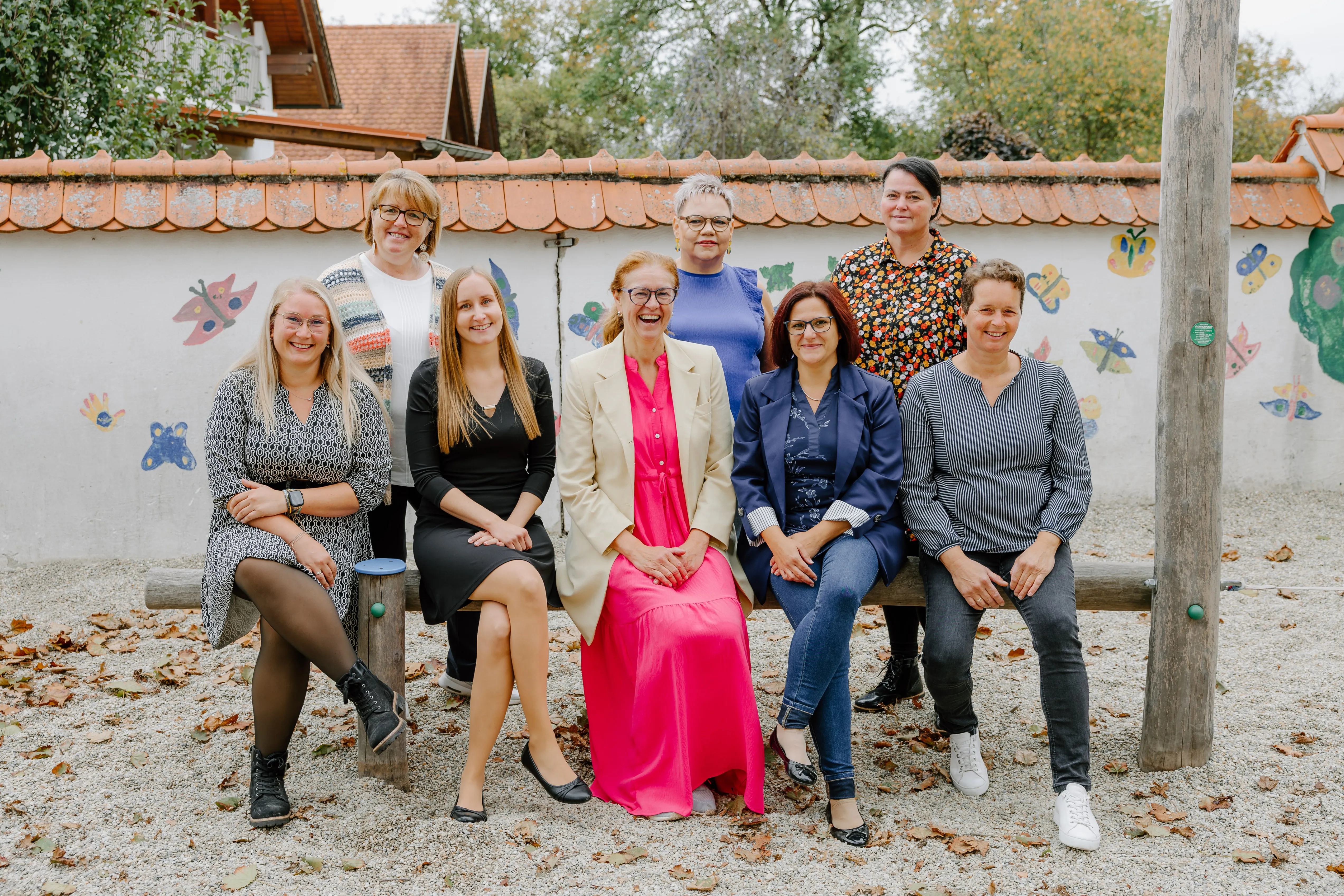 Eine Gruppe von Frauen posiert für ein Foto im Freien. Sie sitzen auf einer Bank und lächeln. Hinter ihnen stehen zwei Frauen.