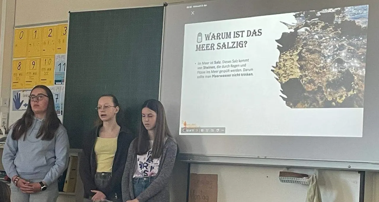 Zwei Frauen stehen in einem Klassenzimmer, wahrscheinlich Studentinnen, eine schaut in die Kamera. Eine Projektionsleinwand zeigt eine Frage, warum Meerwasser salzig ist.
