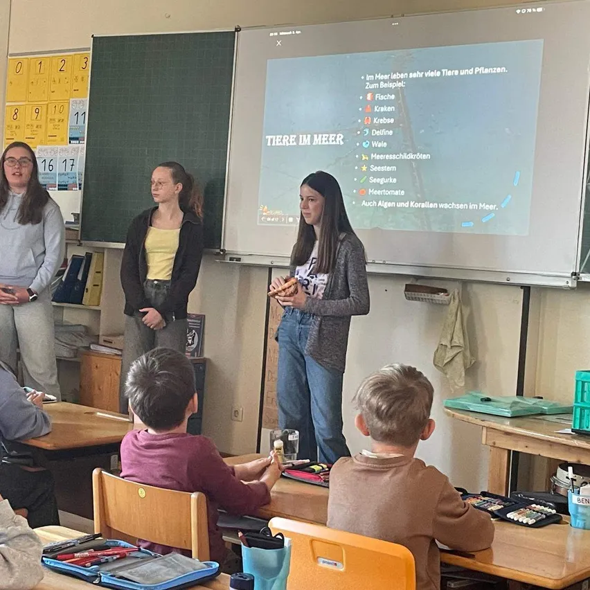 Drei Schüler stehen vor einer Projektionsleinwand, die Informationen über Meerestiere zeigt. Sie sind von Schülern umgeben, die an Tischen in einem Klassenzimmer sitzen.