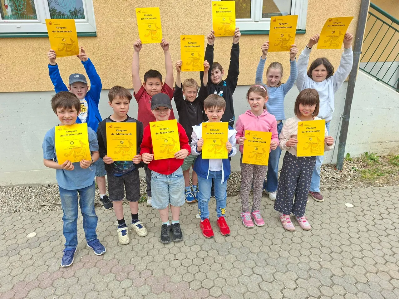 Eine Gruppe von Kindern, die gelbe Zertifikate für Mathematik in der Hand halten, stehen auf einer gepflasterten Fläche, mit einem Gebäude im Hintergrund.