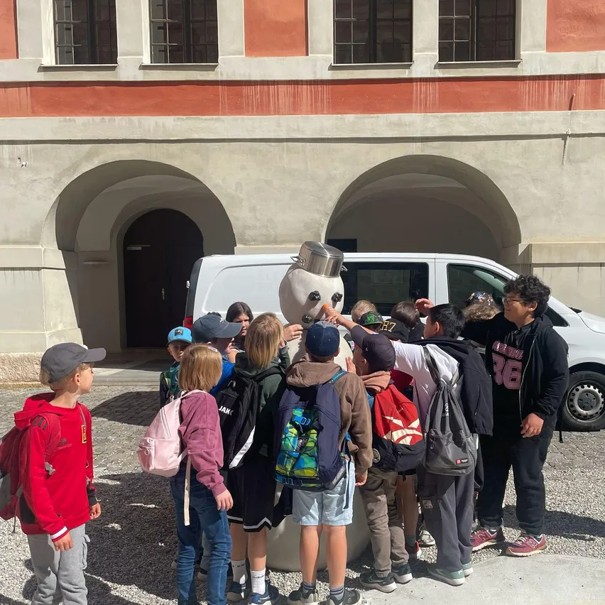 Eine Gruppe von Kindern und ein Mann stehen um einen großen Schneemann vor einem Gebäude. Die Kinder tragen Rucksäcke und Mützen, und der Mann hat einen Bart und eine Brille. Ein Van ist hinter ihnen geparkt.