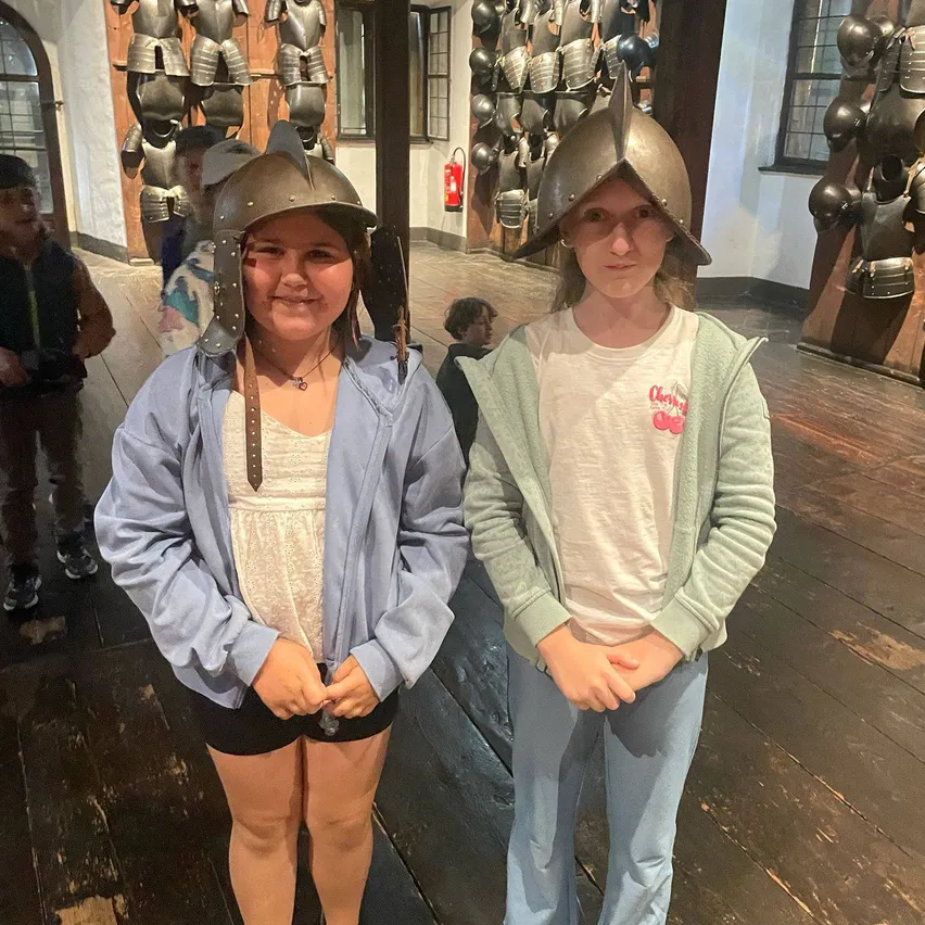 Zwei Mädchen in mittelalterlichen Helmen posieren für ein Foto in einem Museum mit Holzboden und einer Wand voller Rüstungen.