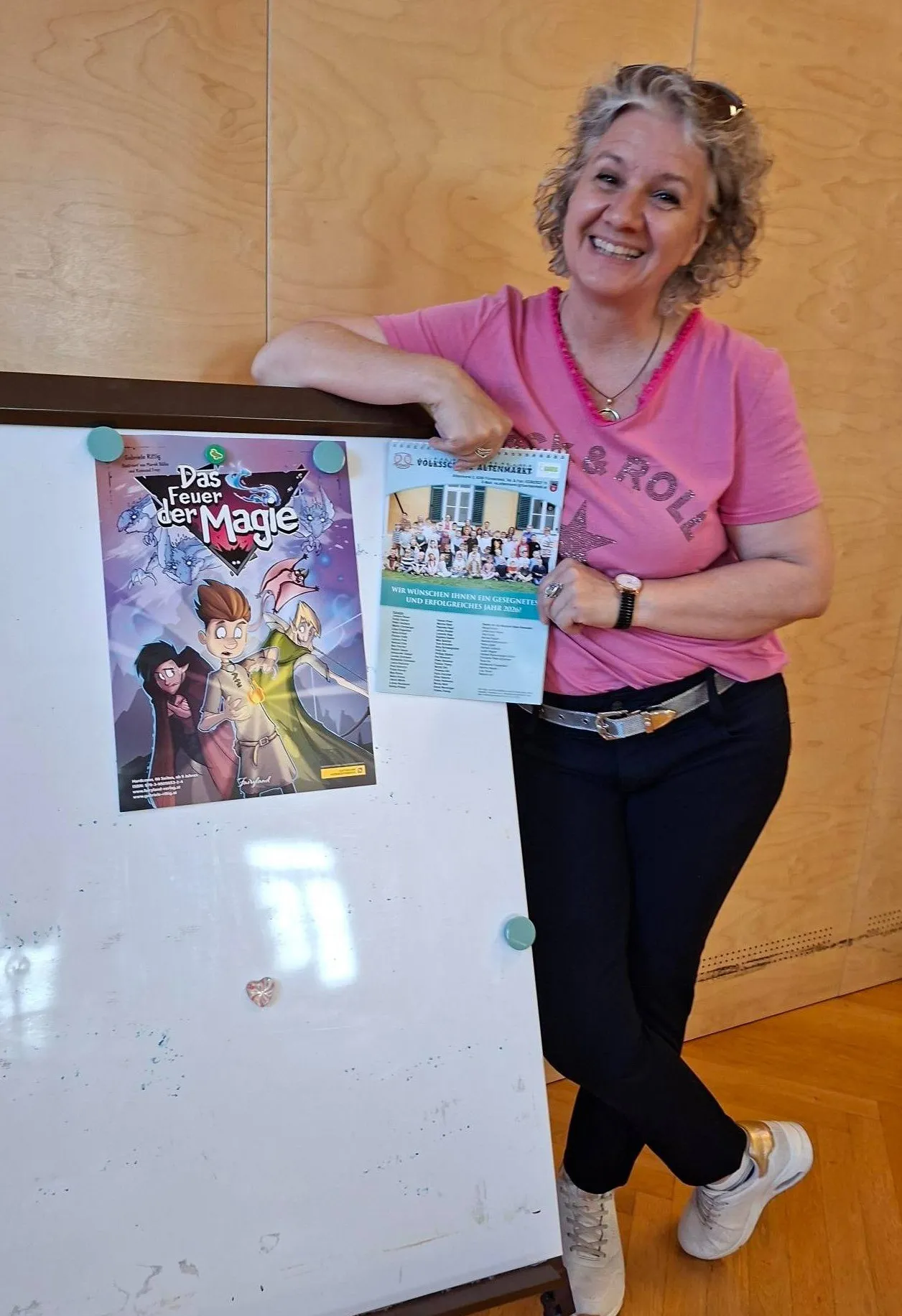 Eine Frau hält ein Buch mit dem Titel 'Das Feuer der Magie' mit einem Schulgruppenfoto, trägt ein rosa Shirt und einen silbernen Gürtel und steht neben einem Whiteboard.