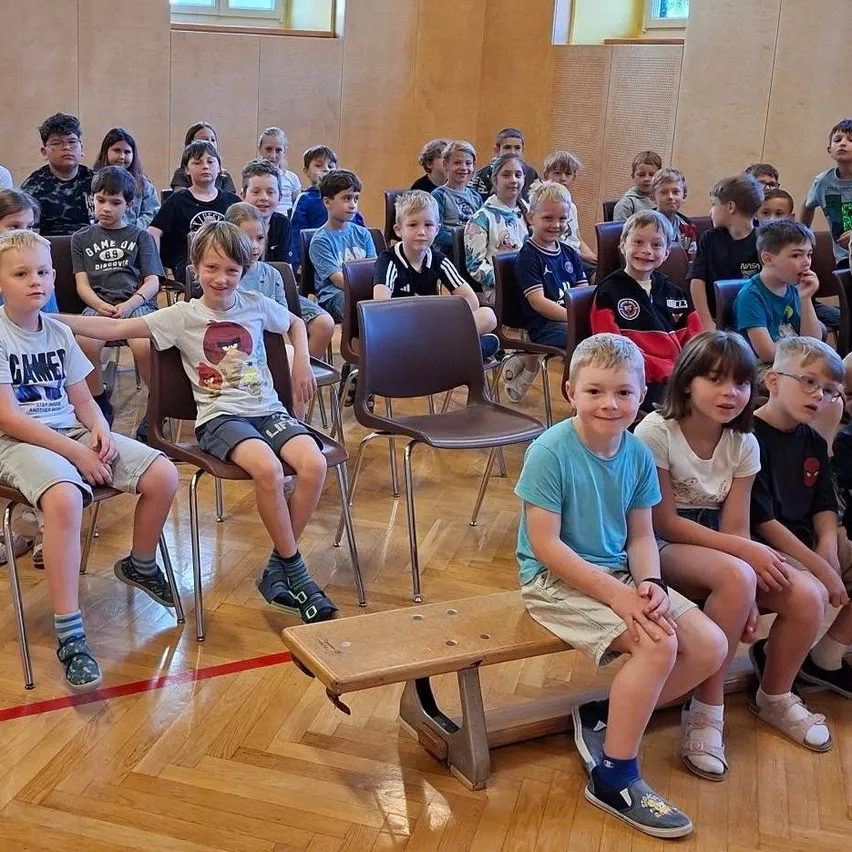 Ein Klassenzimmer voller Kinder, Jungen und Mädchen, die in Stühlen um eine zentrale Bank herum sitzen, wobei einige Brillen und Turnschuhe tragen.