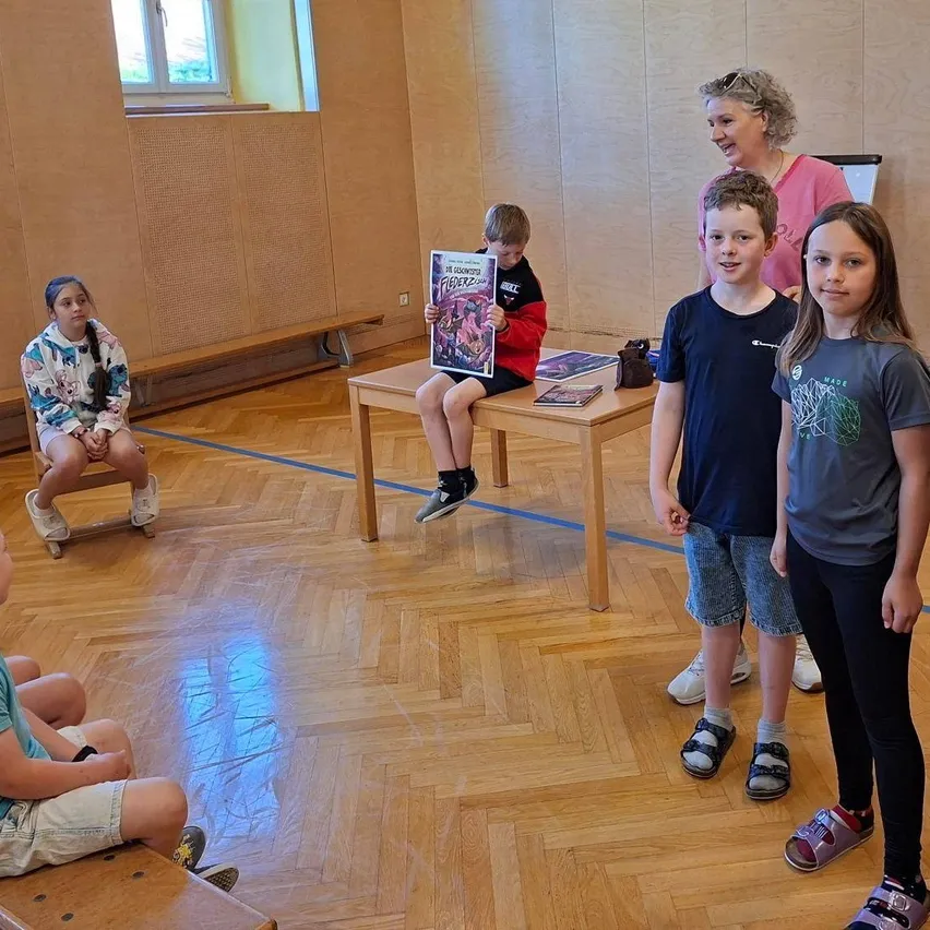 Mehrere Kinder sitzen auf Bänken in einem Raum. Ein Junge sitzt auf einem Tisch und hält ein Comic-Buch. Eine Frau steht hinter dem Tisch.