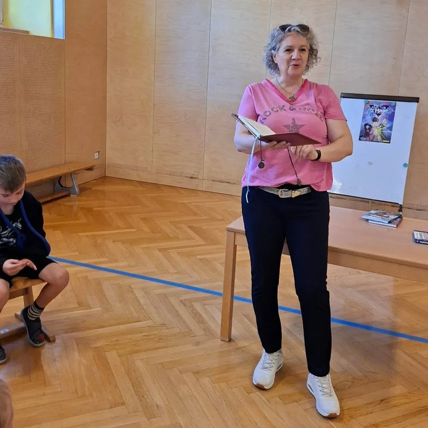 Eine Frau in einem rosa T-Shirt liest in einem Raum ein Buch. Ein Junge sitzt auf einer Bank in der Nähe. Ein Tisch mit Büchern befindet sich rechts und eine Tafel mit einem Poster an der Wand.