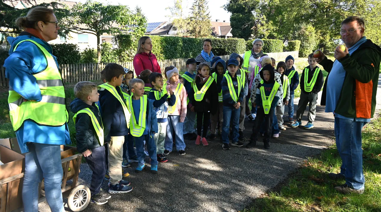 Eine Gruppe von Kindern, die Sicherheitswesten tragen, steht am Straßenrand, mit Erwachsenen im Hintergrund.