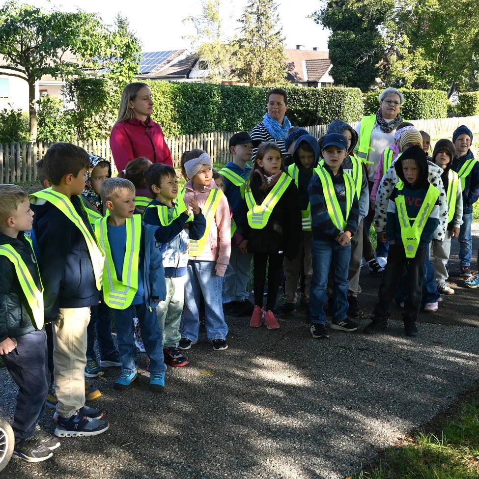 Eine Gruppe von Kindern, die Sicherheitswesten tragen, steht am Straßenrand, mit Erwachsenen im Hintergrund.