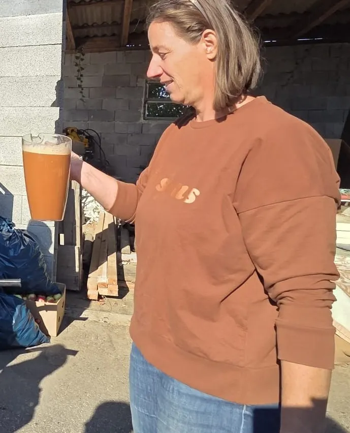 Eine Frau in einem braunen Shirt mit dem Wort 'SUS' darauf, hält ein Glas Orangensaft.