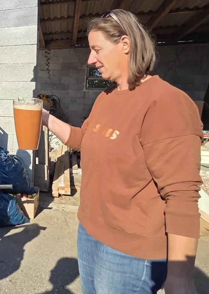 Eine Frau in einem braunen Shirt mit dem Wort 'SUS' darauf, hält ein Glas Orangensaft.