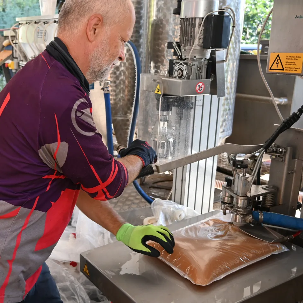 Ein älterer Mann in einem lila und grauen Shirt arbeitet an einer Maschine. Er hält ein Werkzeug und scheint etwas zu reparieren.