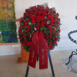 Ein großer Kranz aus roten Rosen mit grünen Blättern und einer roten Schleife mit dem Wort Zum Abschied darauf. Er steht auf einem blauen Ständer auf einem gefliesten Boden.