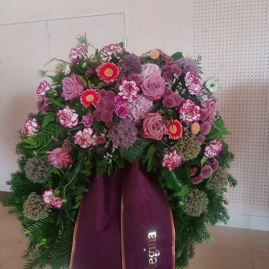 Eine lebhafte Blumenzusammenstellung mit einer Mischung aus rosa, lila und weißen Blumen und grünem Laub steht auf einem gefliesten Boden. Ein lilafarbenes Band mit dem Wort 'Engel' ist befestigt.