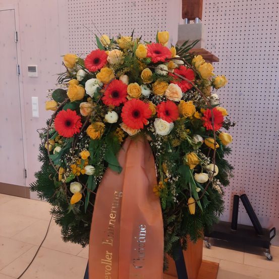 Ein großer Blumenkranz aus roten, gelben und weißen Blumen mit grünen Blättern ist an einer weißen Wand ausgestellt. Ein orangefarbenes Band mit der Aufschrift 'Ehrnerrung' ist um den Kranz gebunden.