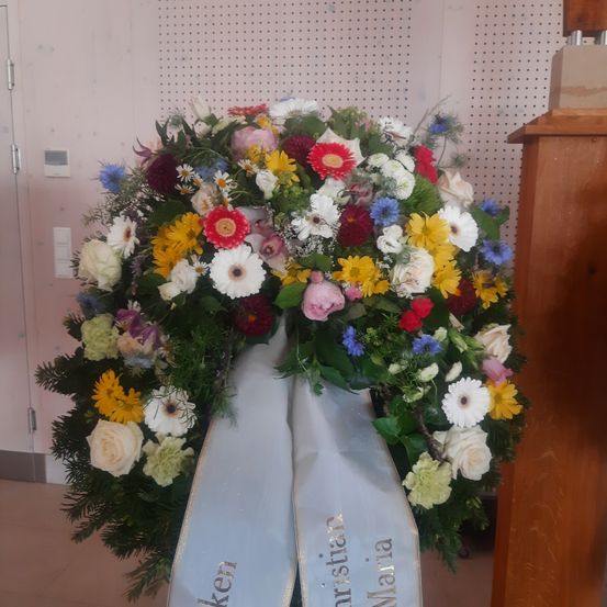 Ein farbenfroher Blumenkranz mit Bändern, die 'Christian' und 'Maria' stehen, ist auf einer Holzoberfläche platziert.