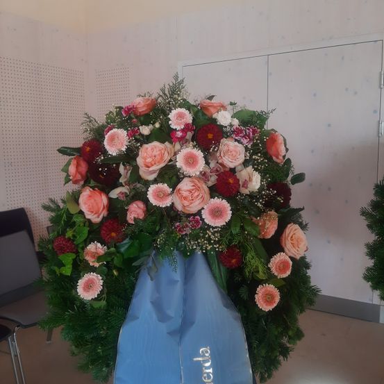 Ein großer Blumenkranz mit verschiedenen rosa und roten Blumen, Grünpflanzen und einer blauen Abdeckung mit dem Wort 'verda' steht in einem Raum.