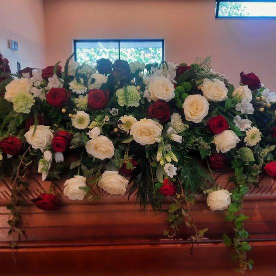 Ein Sarg, geschmückt mit einer aufwendigen Blumendekoration aus weißen und roten Rosen, grünen Blättern und anderen Blumen, in einem Raum mit rosa Wänden und einem Fenster.