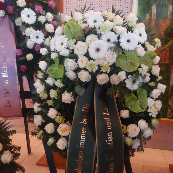 Ein Kranz aus weißen Blumen, einschließlich Gänseblümchen, Rosen und grünen Blättern, wird mit einem schwarzen Band mit der Aufschrift 'danke immer bei Dir' ausgestellt. Dahinter steht ein Banner mit der Aufschrift 'Ilse Helt Michael & Gail.' Die Umgebung scheint ein Innenbereich zu sein.