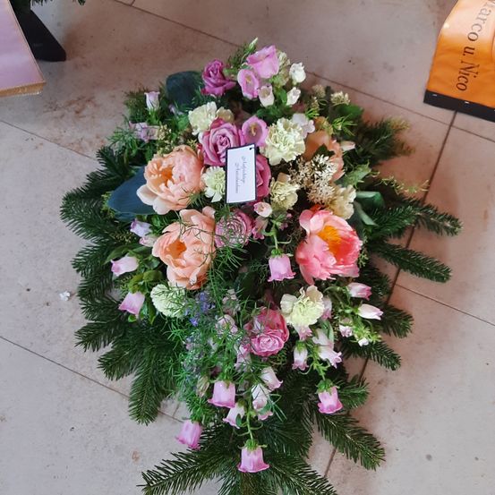 Eine große Blumengestecke aus pinken und weißen Blumen mit Grün, möglicherweise für einen Gedenkgottesdienst. Es hat eine kleine Karte mit Text befestigt.