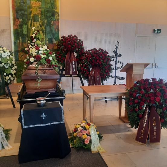 Eine Beerdigungseinrichtung mit zwei Särgen, Blumenarrangements, einem Tisch und einem Podium in einem Raum. Ein Kreuz steht auf einem Tisch neben den Särgen.