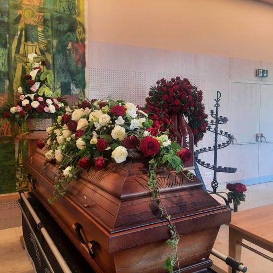 Ein hölzerner Sarg ist mit roten und weißen Blumen in einem Raum mit einem großen Gemälde und einem Ausgangsschild geschmückt.