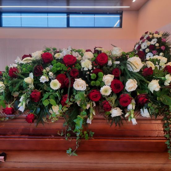 Ein großer Blumenstrauß mit roten, weißen und grünen Blumen schmückt einen Sarg. Der Raum hat eine weiße Wand und ein Fenster mit hellem Licht.