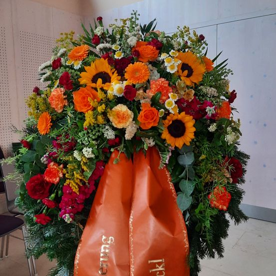 Ein großes Blumenarrangement mit einer Vielzahl von Blumen, darunter rote, orange und gelbe Blüten, Sonnenblumen und Grünpflanzen, gebunden mit einem orangefarbenen Band, das 'Sympathie' und 'Trauer' steht.