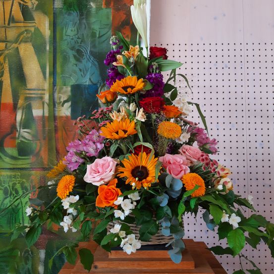 Eine farbenfrohe Blumenarrangement in einem Korb, bestehend aus Sonnenblumen, Rosen und anderen bunten Blumen, steht auf einem Holztisch vor einer farbenfrohen Wand mit einem gepunkteten Muster.