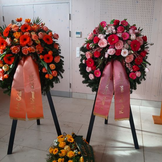 Zwei Kränze mit Bändern stehen auf einem Boden mit weißen Fliesen. Ein Kranz hat rote, rosa und weiße Blumen, während der andere orange und gelbe Blumen hat. Beide Kränze haben Bänder mit goldenen Buchstaben. Der Hintergrund hat eine weiße Wand mit kleinen Löchern.