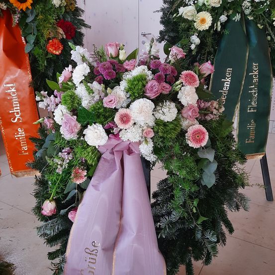 Ein Nahaufnahme einer Blumenarrangements mit weißen, rosa und lila Blumen. Eine lila Schleife ist um den Strauß gebunden. Die Blumen sind von grünen Blättern umgeben.