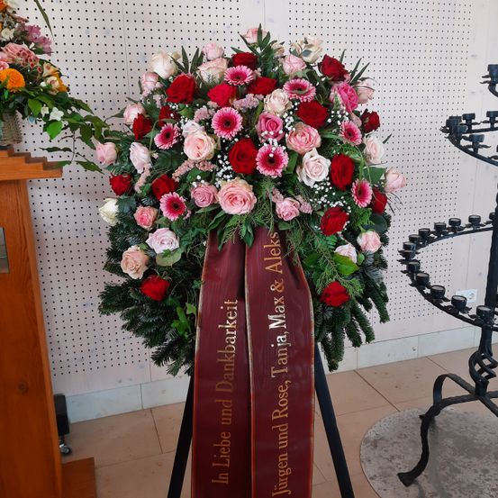 Ein großer Blumenstrauß mit roten, rosa und weißen Rosen sowie Babys Atmung wird an einer weißen Lochwand ausgestellt. Ein rotes Band mit Goldtext ist befestigt.