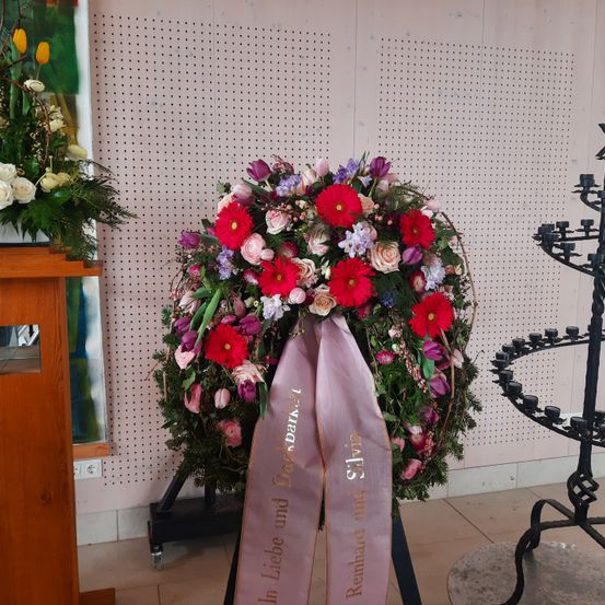 Ein Kranz mit rosa Schleifen, die 'In Gedenken an Silvia' und 'Reinhard und' sagen, geschmückt mit Blumen, steht in einem Raum mit gefliestem Boden.